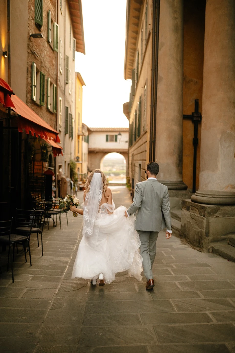 Bride and groom walking hand in hand down a narrow cobblestone street with historic buildings on either side.