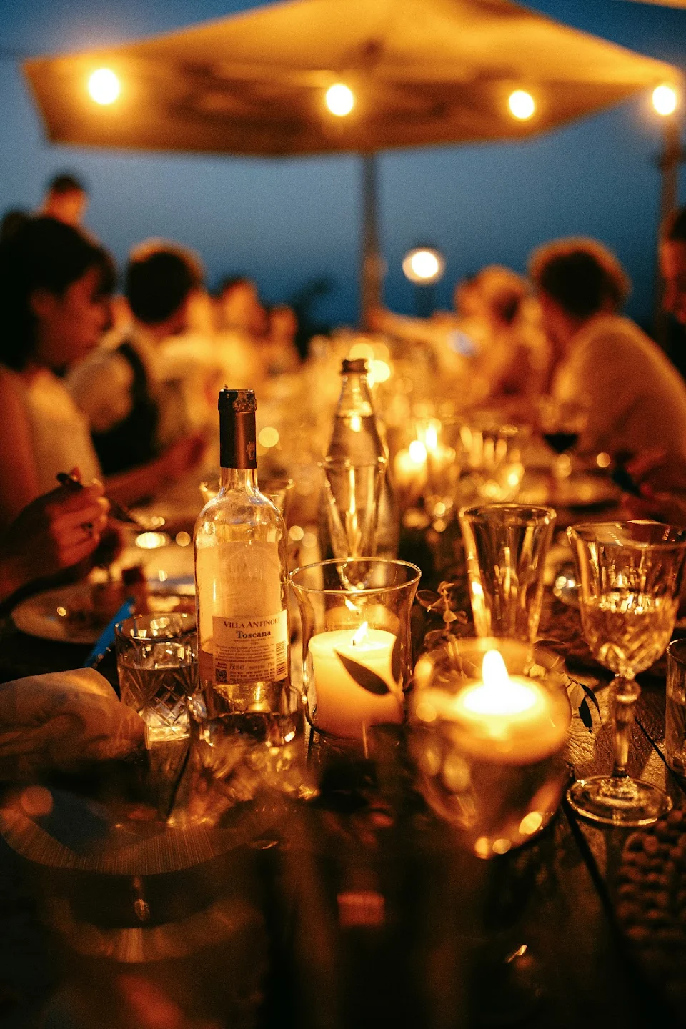 Evening outdoor dinner party with lit candles, wine bottle, and guests dining under string lights.