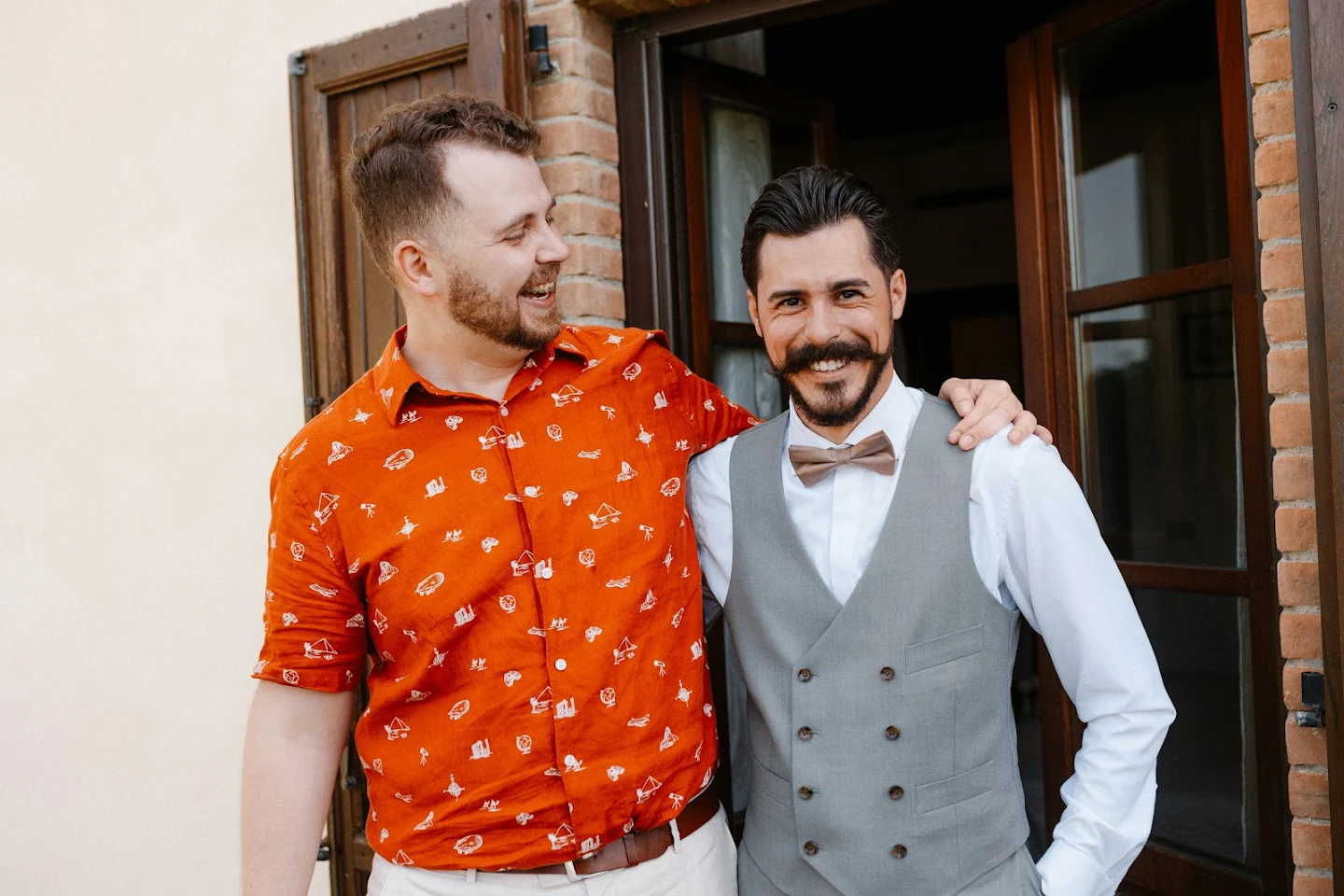 Two men smiling, one wearing an orange patterned shirt and the other in a grey vest with a brown bow tie, standing outside a wooden door.