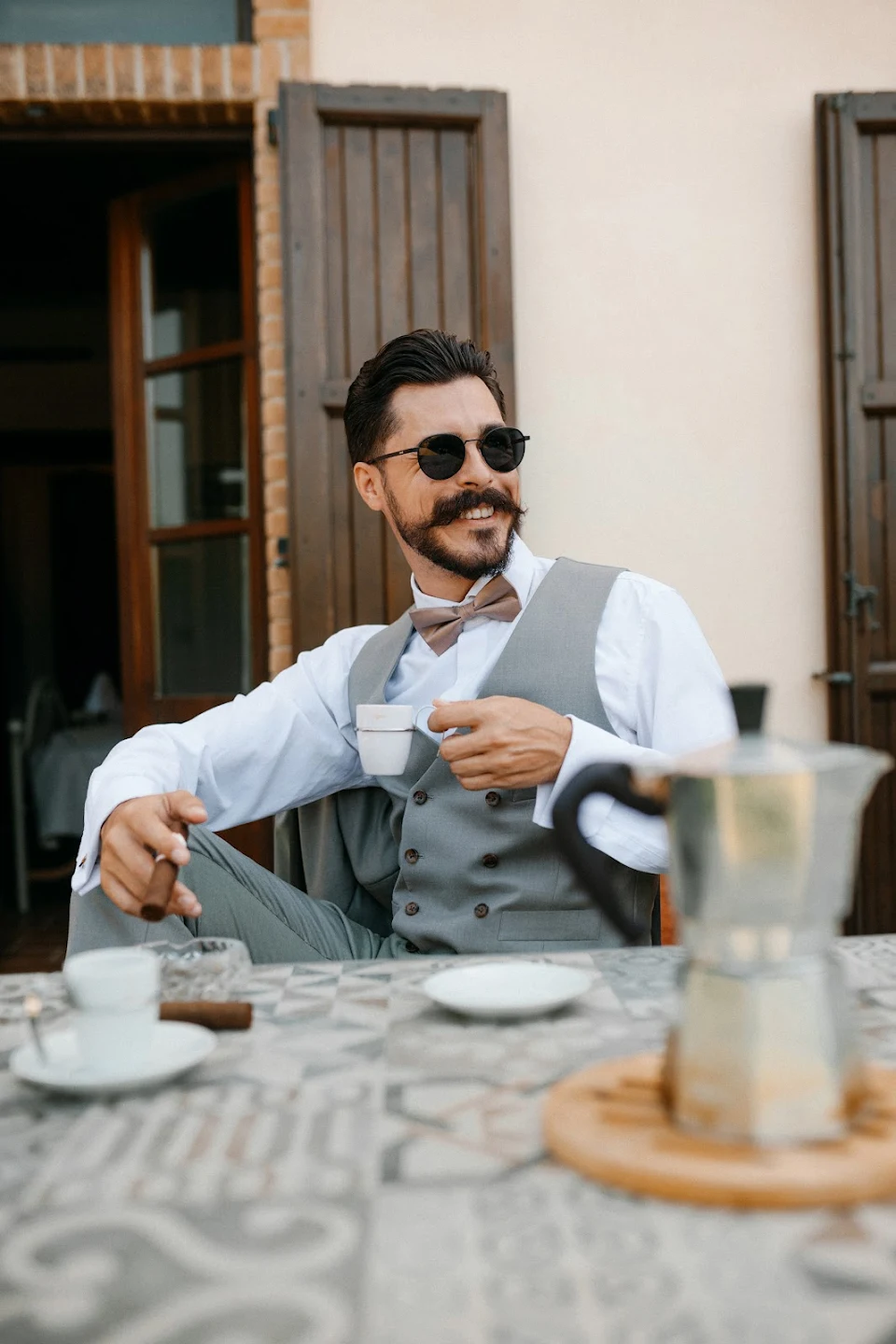 Smiling man in sunglasses and a gray vest holding a cup of coffee and a cigar while sitting at a table outside.