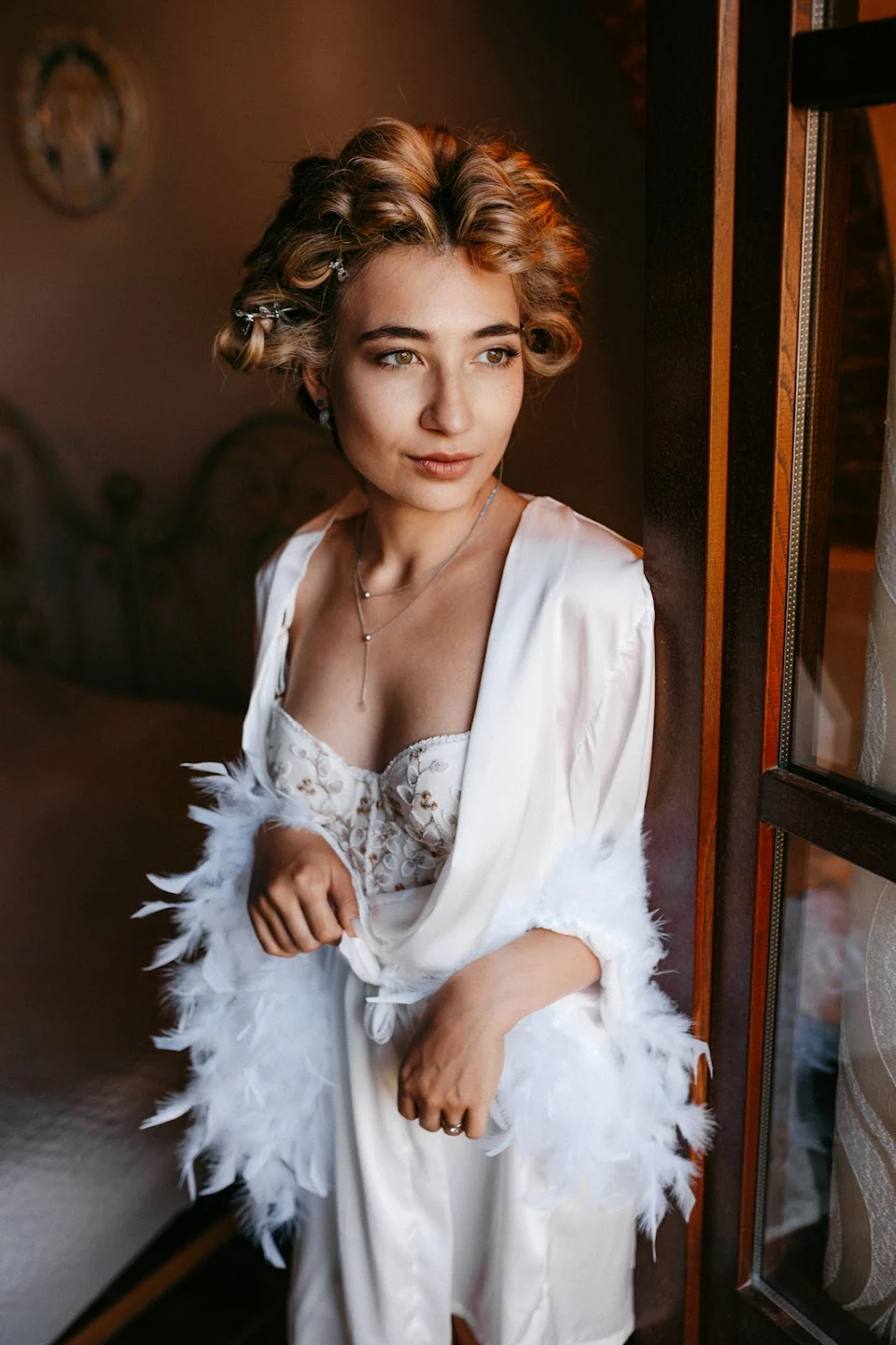Young woman with curled hair clips wearing a white robe with feathered sleeves and a detailed corset, standing by a window.