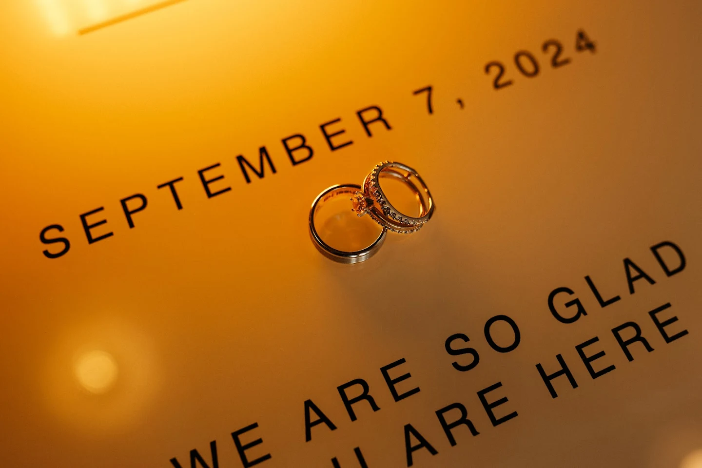 Two gold wedding rings placed on a card with the date September 7, 2024, and the text 'WE ARE SO GLAD YOU ARE HERE' in black letters.
