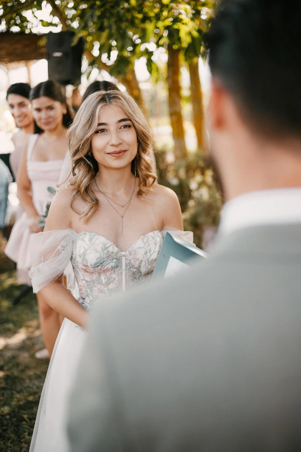 Bride with wavy blonde hair and an off-shoulder floral wedding dress smiling during an outdoor ceremony, with bridesmaids in soft pink dresses in the background.