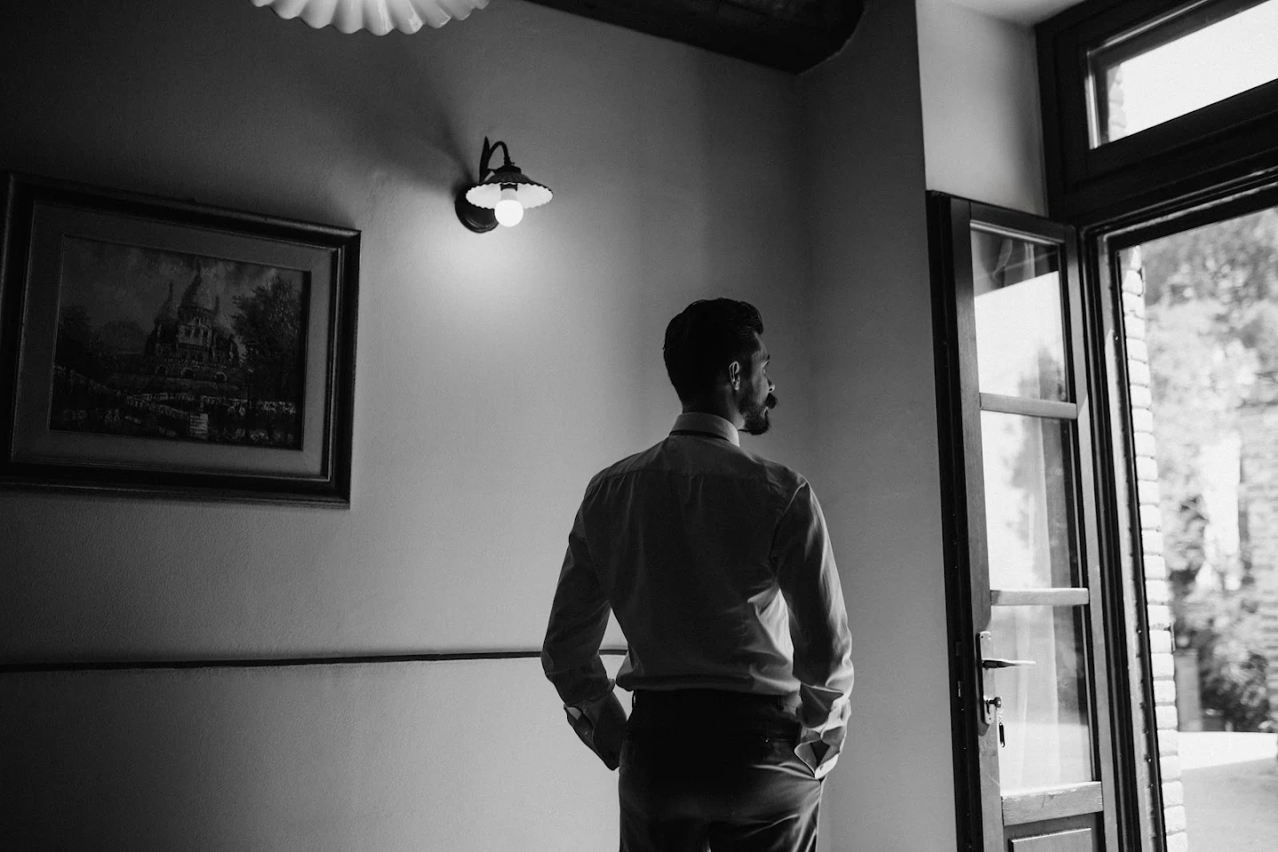 Man in dress shirt and pants standing with hands in pockets, looking out an open door with light coming in.