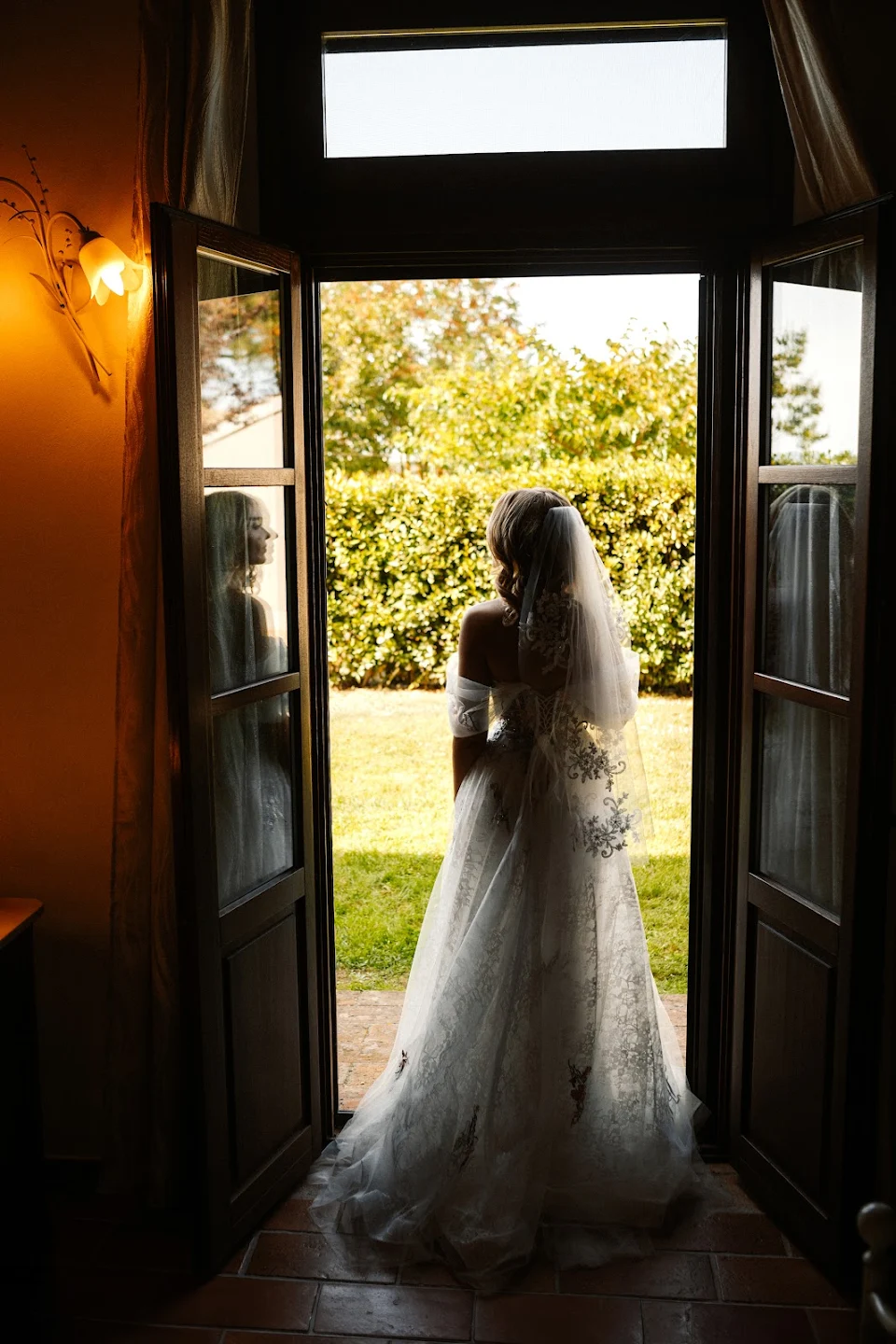 Bride in a lace wedding dress and veil standing in an open doorway looking out to a sunlit garden.