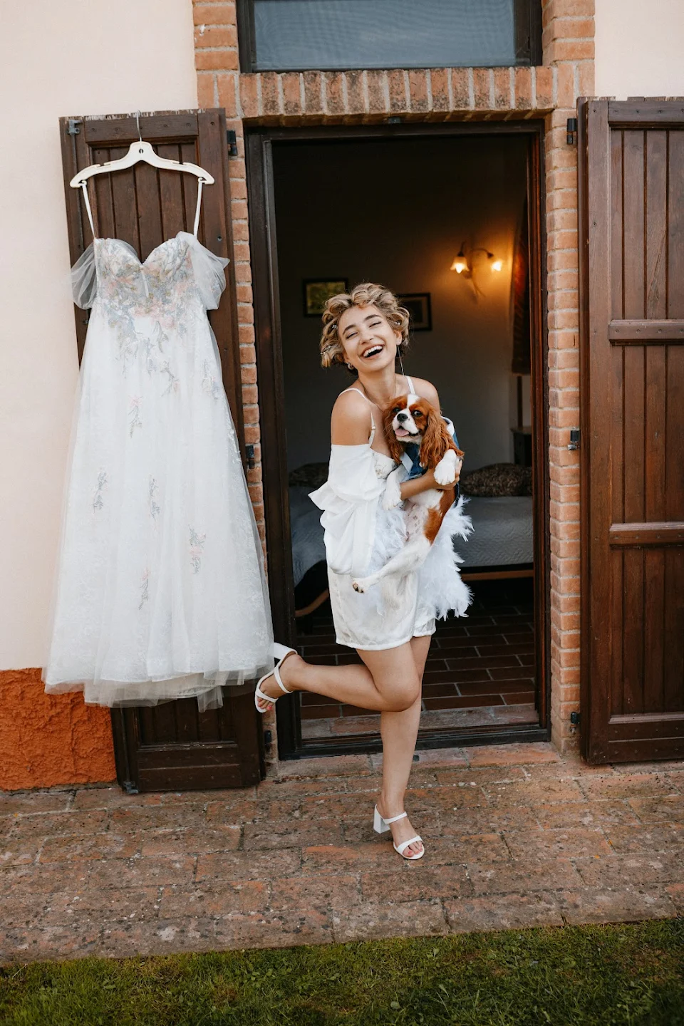 Happy bride in white robe holding a small dog outside a doorway with her wedding dress hanging on a wooden shutter.