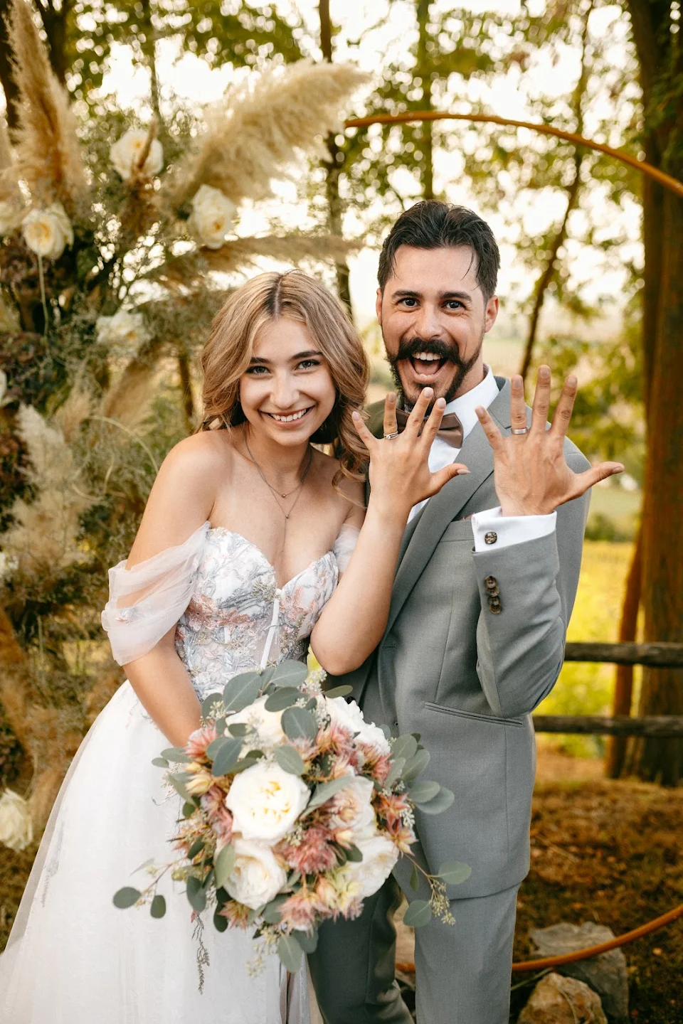Smiling bride and groom showing their wedding rings outdoors with floral decorations in the background.