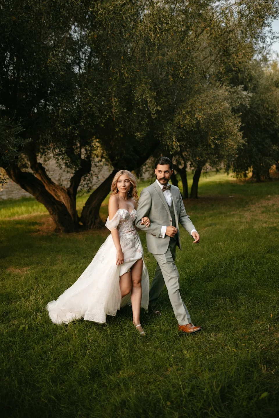 Bride in a white wedding dress with a thigh-high slit walking arm in arm with groom in a light gray suit on green grass near trees.