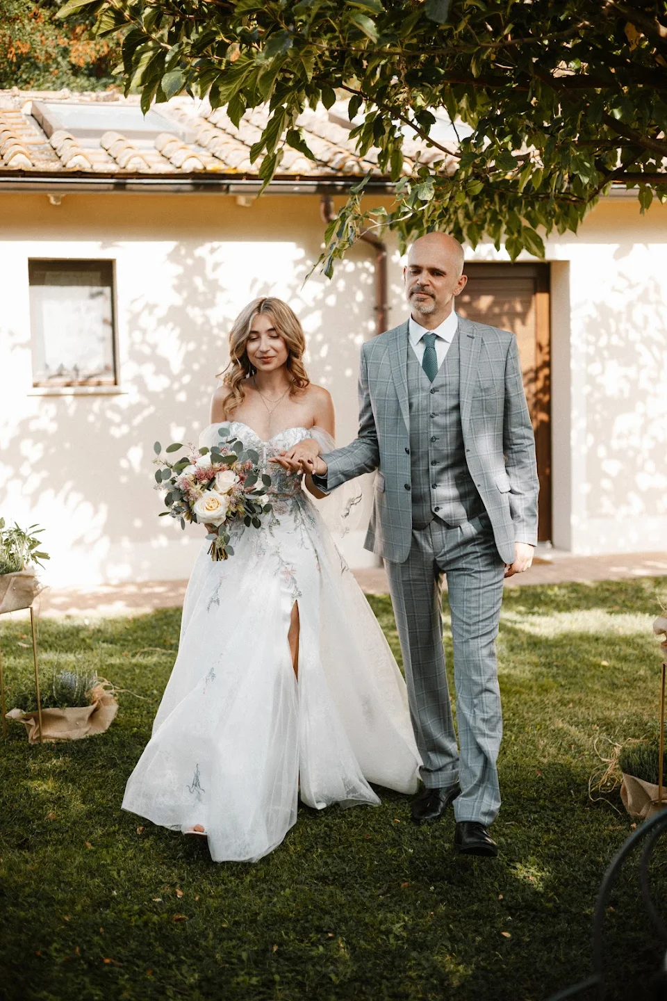 Bride in a white wedding dress holding a bouquet walks hand in hand outdoors with a man in a gray checkered suit and green tie.