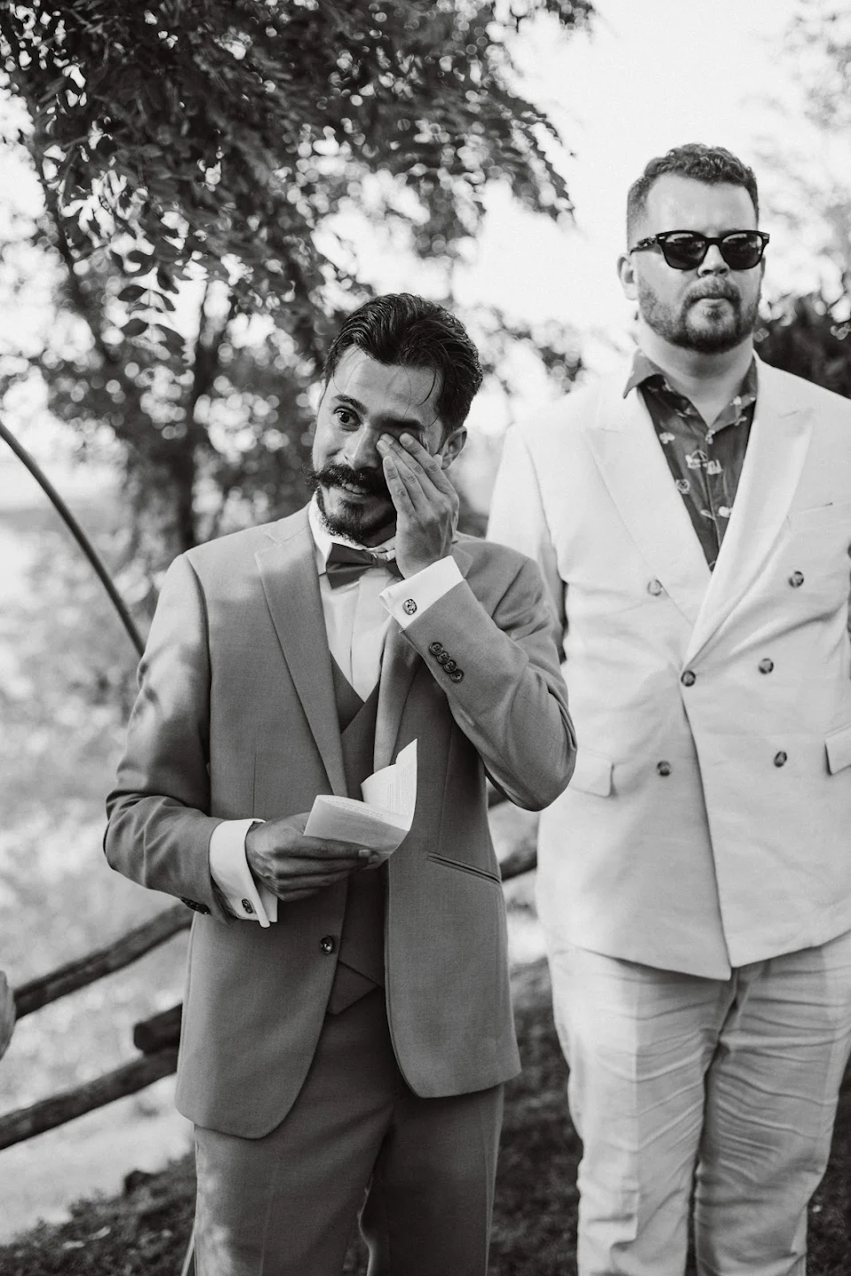 Emotional groom wiping tears while holding a paper, standing next to a man in sunglasses and a light-colored suit outdoors.