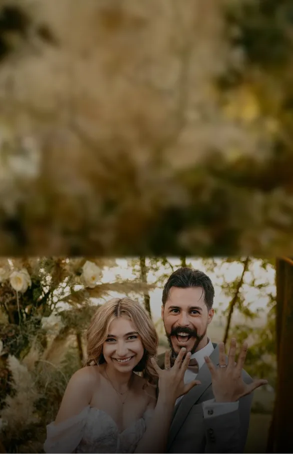 Smiling newlywed couple showing their wedding rings with hands raised outdoors surrounded by greenery and flowers.