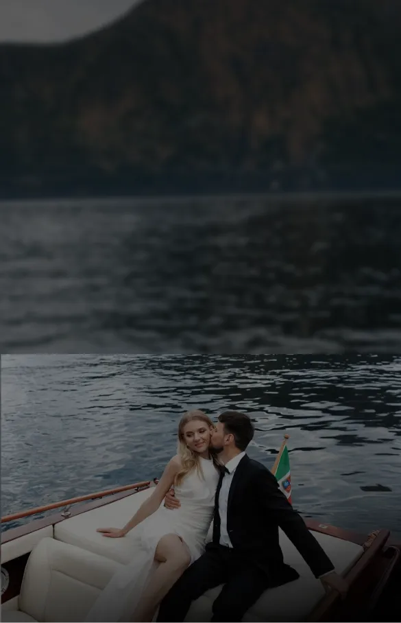 Couple dressed in formal attire sitting on a boat with a lake and mountains in the background; the man is kissing the woman on the cheek.