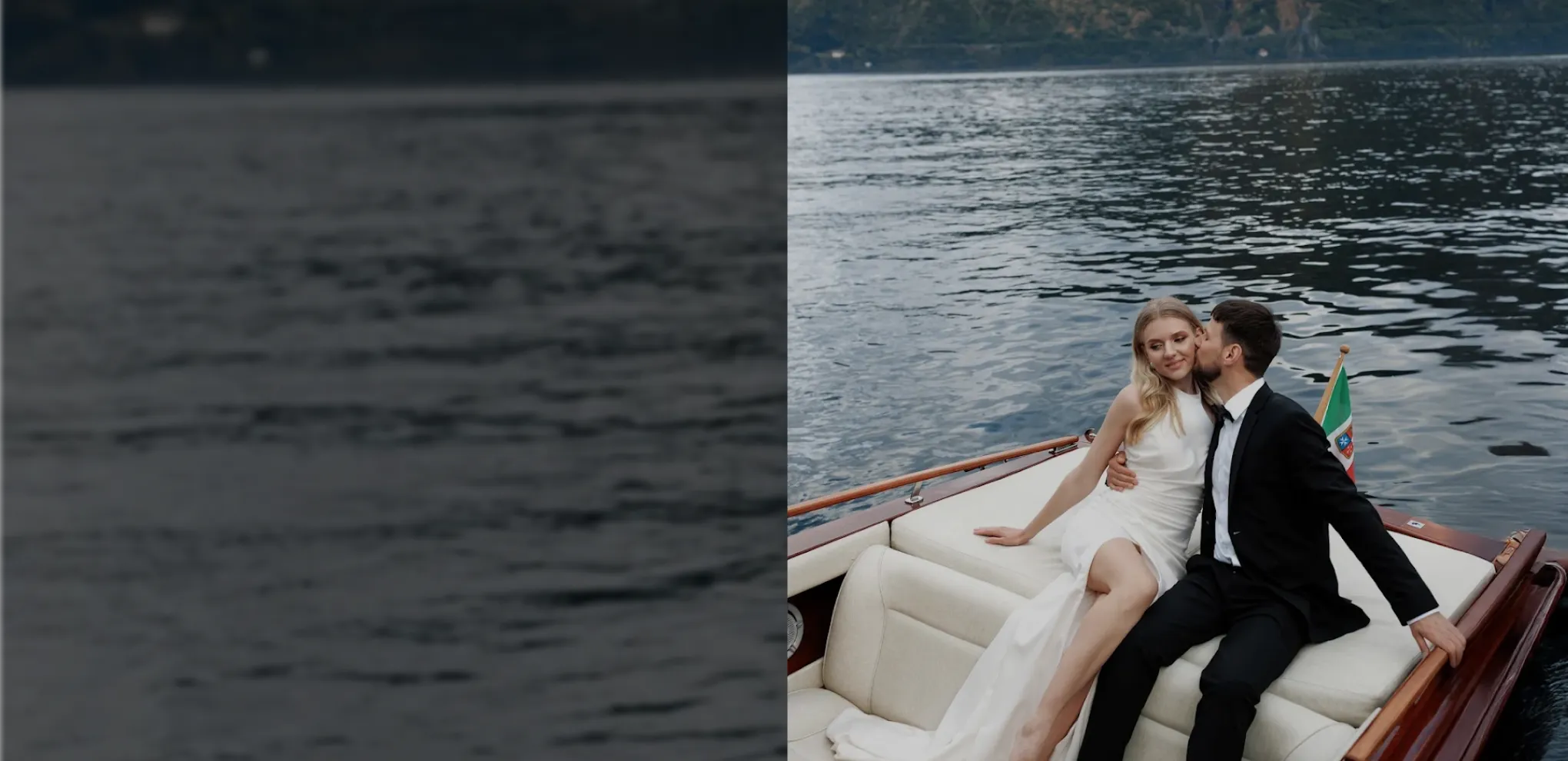 Couple dressed elegantly sitting on a boat with water and mountains in the background, man kissing woman's cheek.