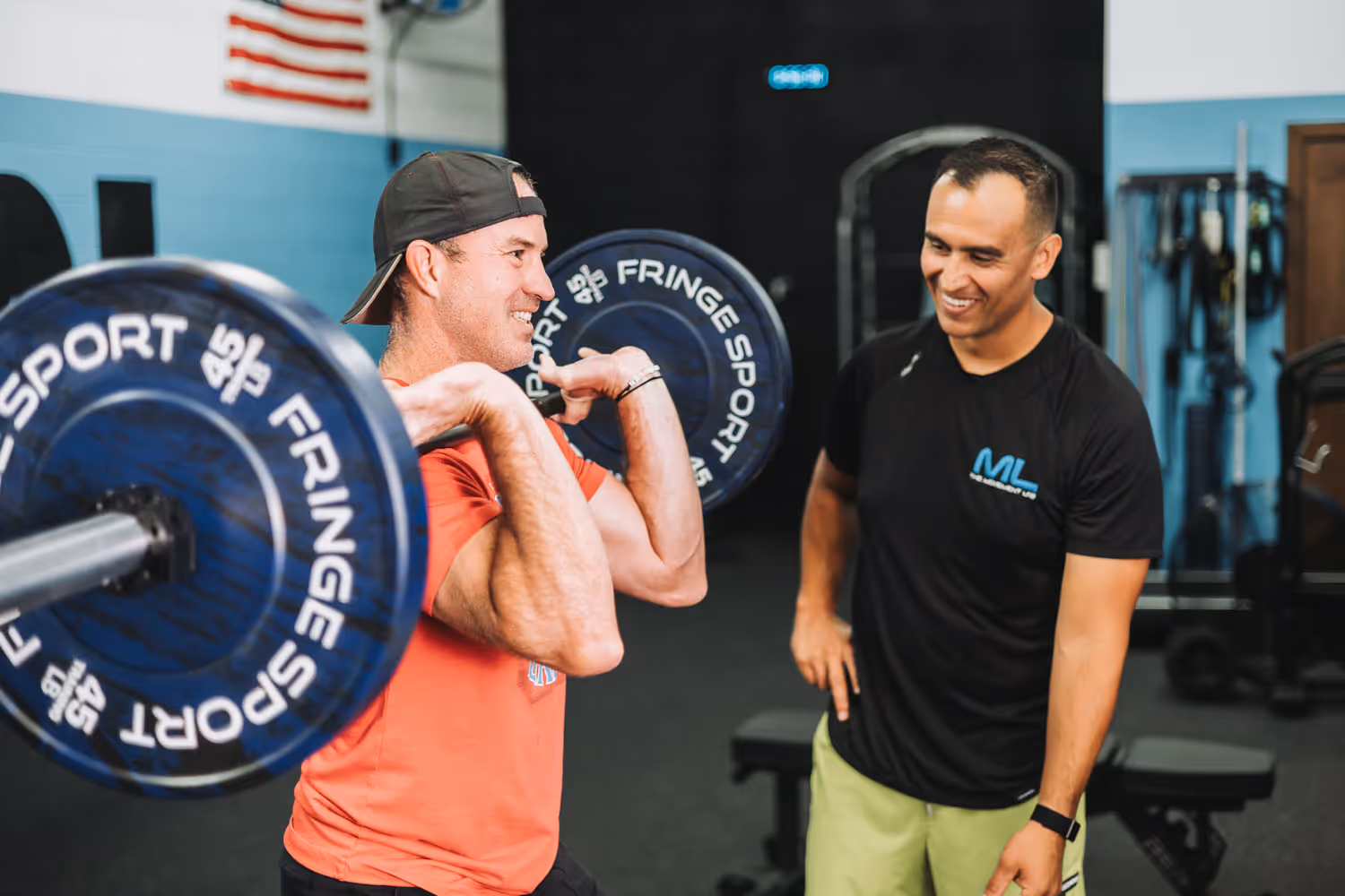 A man lifting a barbell with weights while another man in a black shirt with ML logo watches and smiles inside a gym.