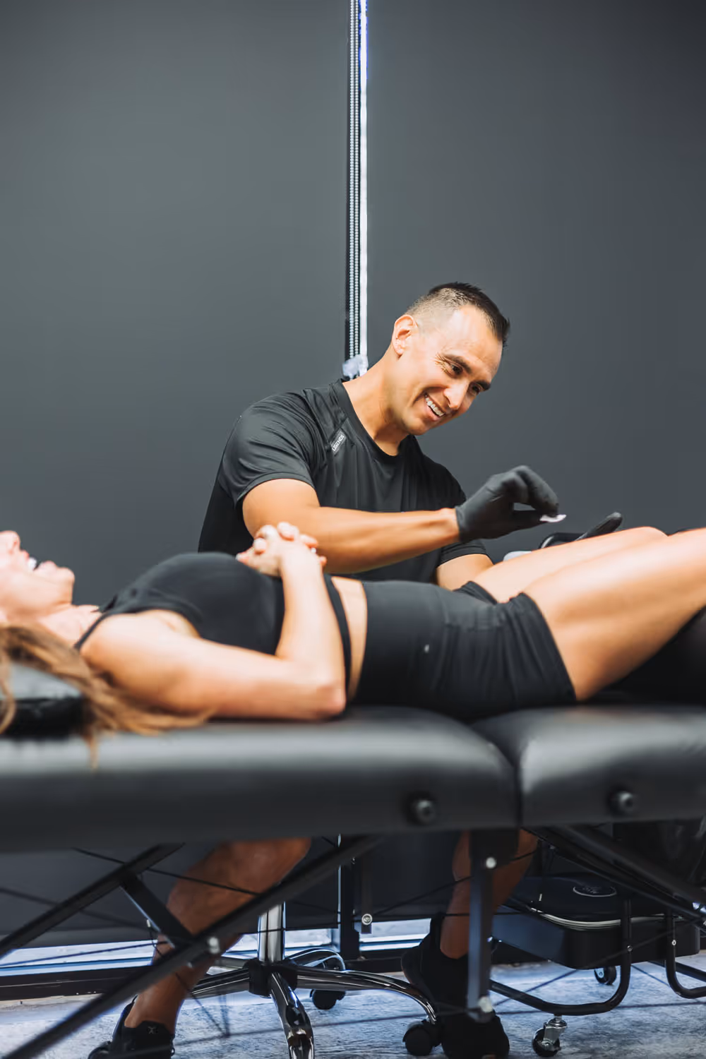 Male therapist wearing gloves preparing to perform a treatment on a woman lying on a black massage table.