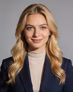 Smiling young woman with long blonde hair wearing a navy blazer and beige turtleneck against a gray background.
