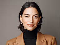 Portrait of a woman with short dark hair wearing a brown blazer and black turtleneck against a gray background.