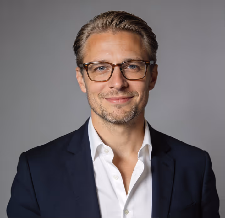 Smiling man with light brown hair and glasses wearing a white shirt and dark blazer against a gray background.