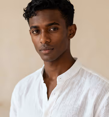 Portrait of a young man with dark curly hair wearing a white collared shirt against a beige background.