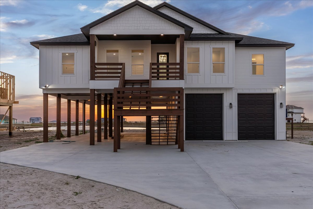 Elevated modern beach house with white siding, dark garage doors, wooden posts, and a central staircase at sunset.