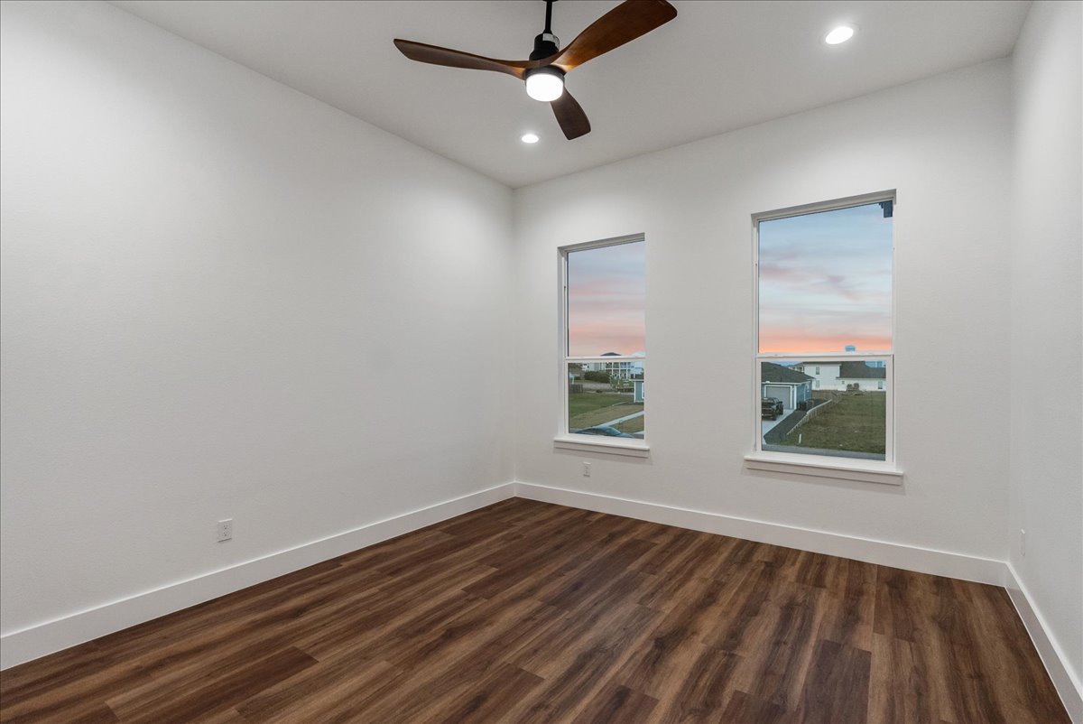 Empty room with white walls, wood flooring, two windows showing a sunset, and a ceiling fan with a light.