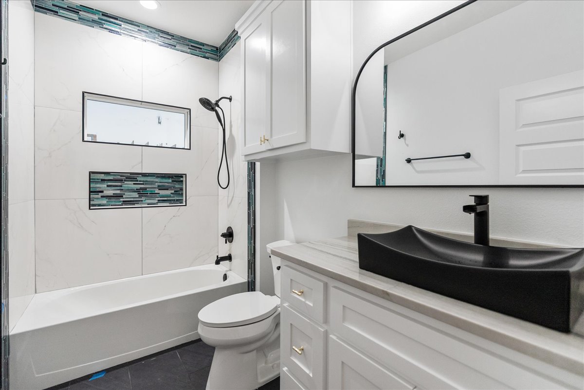 Modern bathroom with white tub, black shower fixtures, decorative tile accent, white cabinetry, and black rectangular vessel sink under a large mirror.