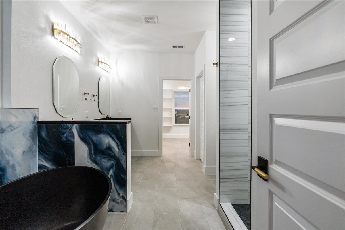 Modern bathroom with black bathtub, dark swirling marble vanity, two mirrors, and glass shower with white tile walls.
