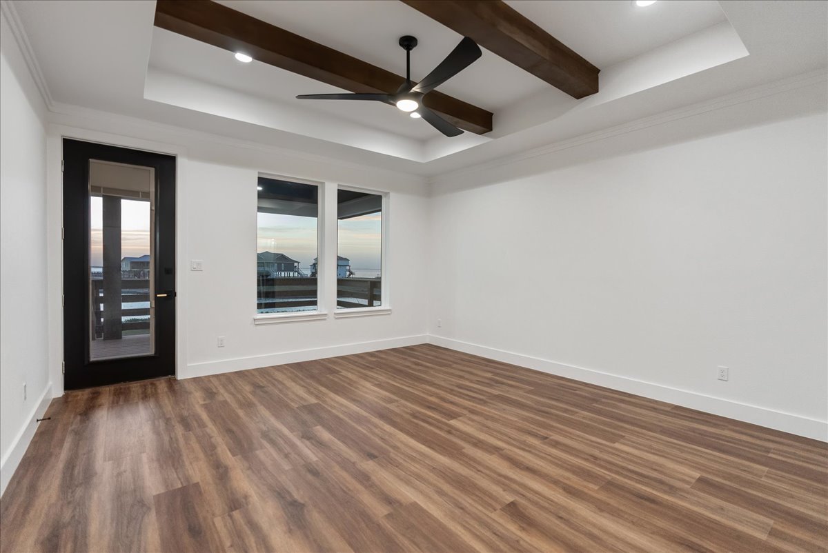 Empty room with wood flooring, white walls, ceiling fan with lights, exposed wooden ceiling beams, black framed glass door, and two windows showing waterfront houses outside.