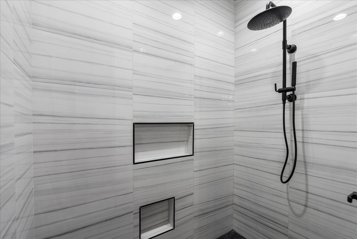 Modern shower area with horizontal striped gray tiles, built-in shelves, and a black rainfall showerhead with handheld attachment.