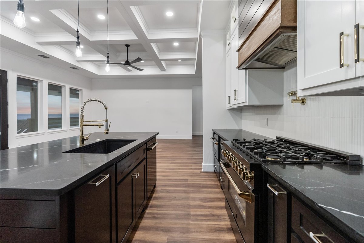 Modern kitchen with dark island featuring a black sink and brass faucet, black stove with brass knobs, white upper cabinets, and wood flooring under recessed ceiling lights and hanging bulbs.