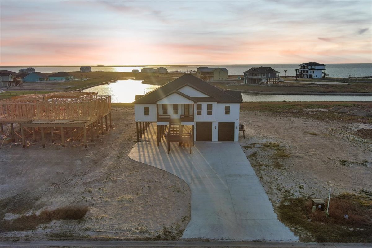 Stilted white house with double garage by a waterfront at sunset, with neighboring houses and a wooden construction frame nearby.