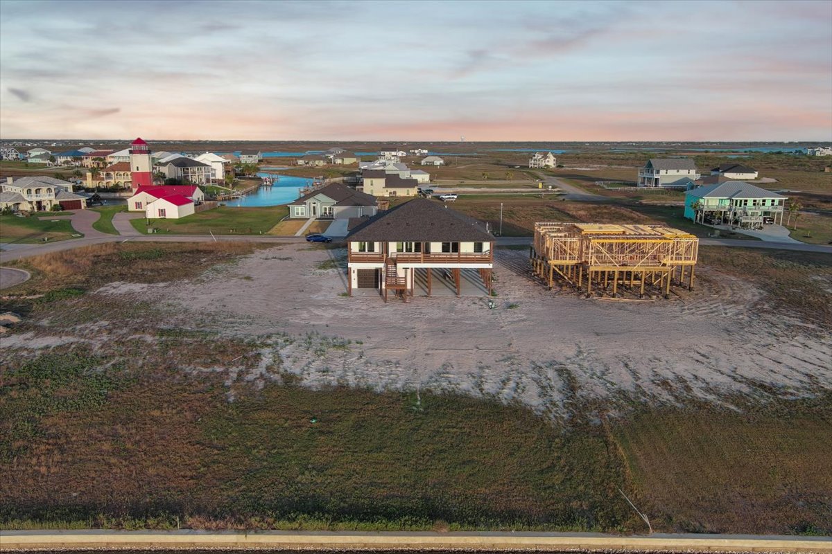 Aerial view of two beachfront houses, one fully built on stilts and the other under construction, with more houses and water channels in the background.