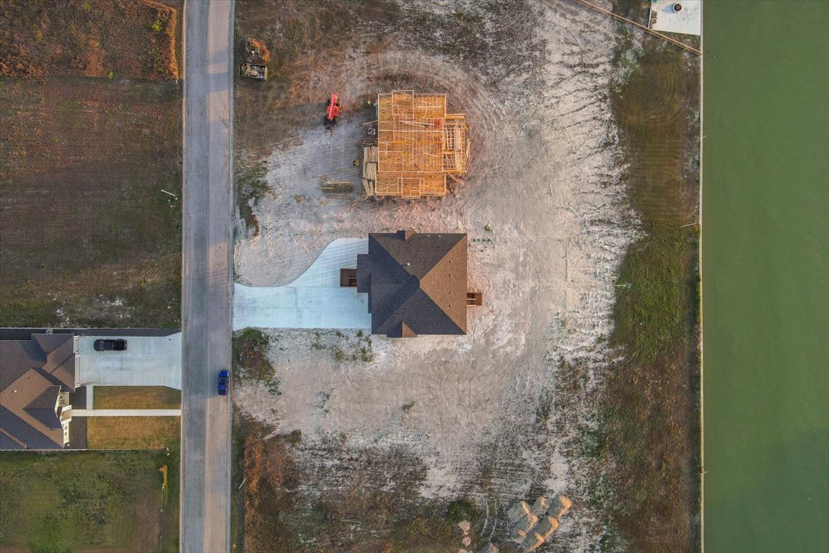 Aerial view of a residential area showing one fully built house with a driveway and one house under construction on a large dirt lot adjacent to a green body of water.