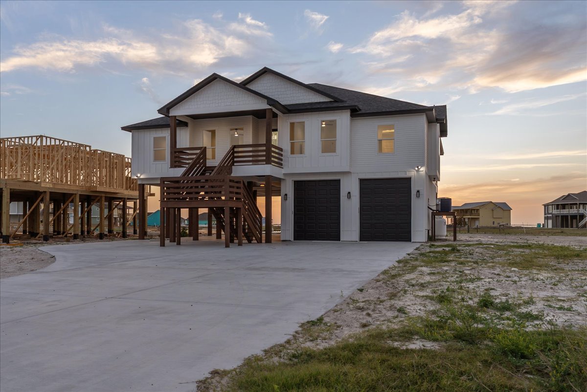 Modern elevated house with two black garage doors, wooden staircases, and a large concrete driveway at sunset.