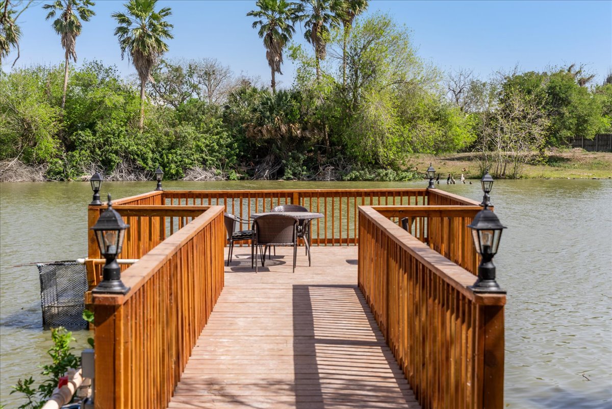 Wooden dock with railing extending over a body of water, featuring a small table and four chairs at the end, surrounded by green trees and clear sky.