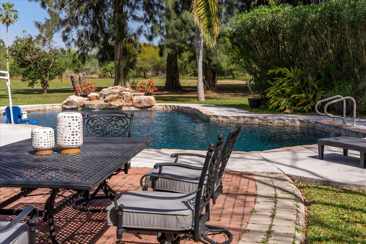 Outdoor patio with black metal table and cushioned chairs next to a curved swimming pool surrounded by trees and greenery.