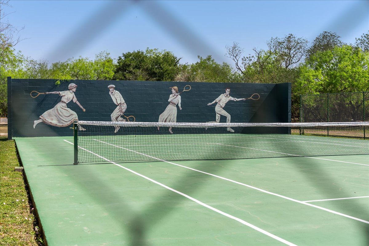 Outdoor tennis court with a green surface and a net, featuring a mural of four vintage-style tennis players painted on a dark wall at the far end.