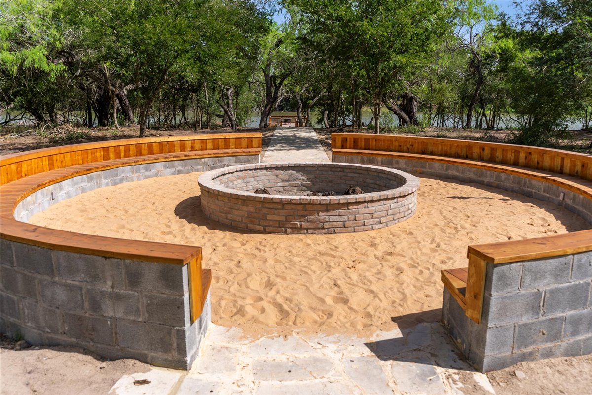 Circular outdoor fire pit surrounded by curved wooden benches on a sandy ground with trees in the background.