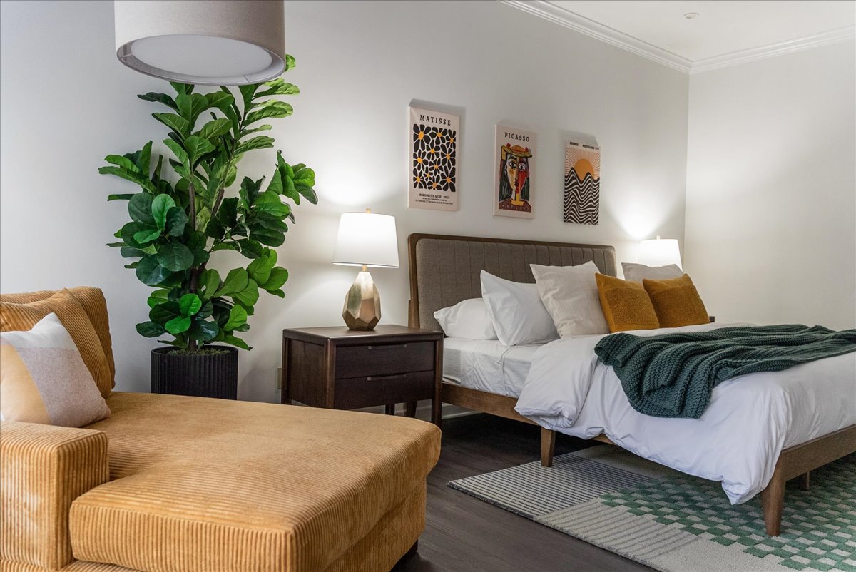 Modern bedroom with a beige upholstered bed, white and mustard pillows, green throw blanket, striped rug, bedside tables with lamps, large green plant, and mustard corduroy chaise lounge.