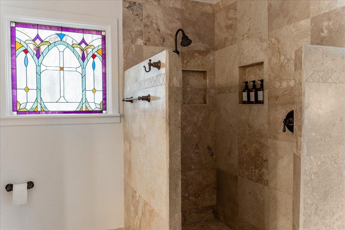 Stone-tiled walk-in shower with built-in shelves holding bottles and a decorative stained glass window on the adjacent wall.