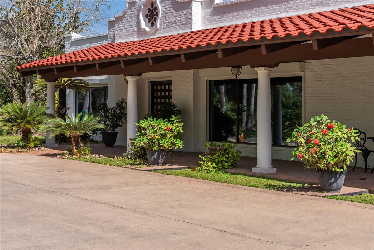 White brick building with red tiled roof, columns, potted plants with red flowers, and a clear blue sky.