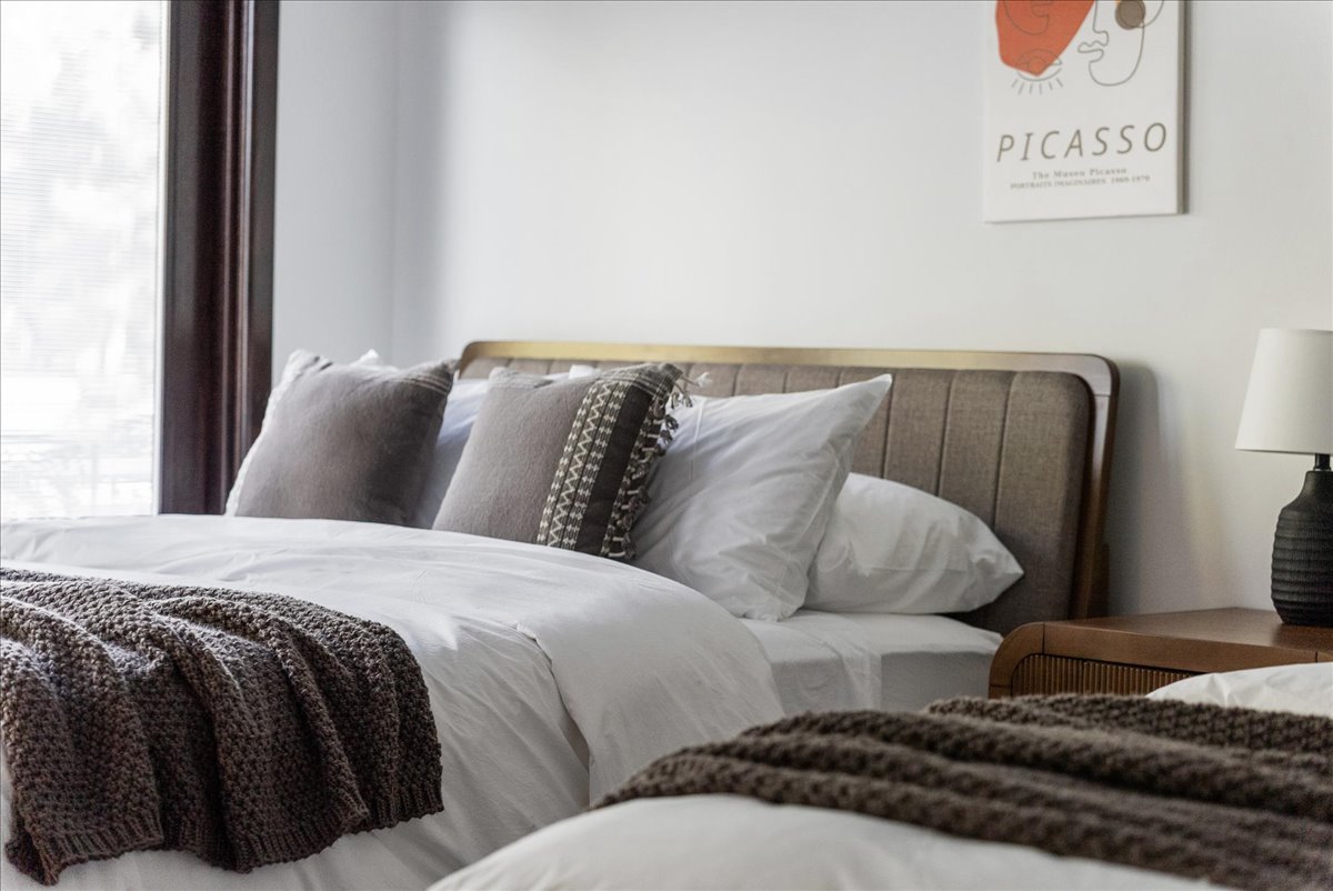 Bedroom with a bed featuring white bedding, brown knitted throw blanket, and decorative pillows, beside a wooden nightstand with a lamp and a Picasso poster on the wall.