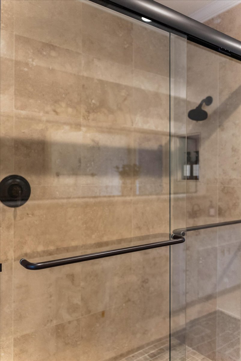 Modern bathroom shower area with beige tiled walls, glass sliding doors, and black metal handles and fixtures.