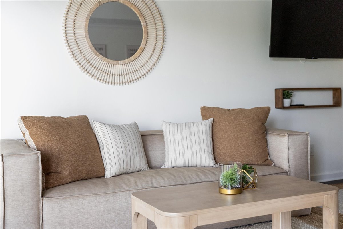 Modern living room with beige sofa adorned with brown and striped cushions, a wooden coffee table with small succulent plants, a round decorative wall mirror, and a wall-mounted TV.