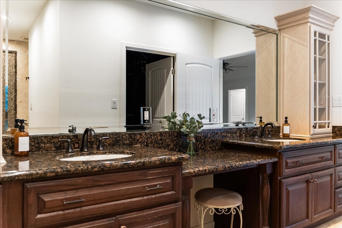 Modern bathroom vanity with two sinks, brown granite countertops, wooden cabinets, a large mirror, and a small stool under the counter.