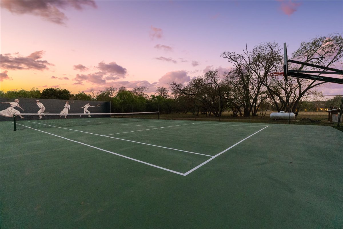 Empty outdoor tennis court with a sunset sky, trees in the background, and painted vintage-style tennis players on a back wall.