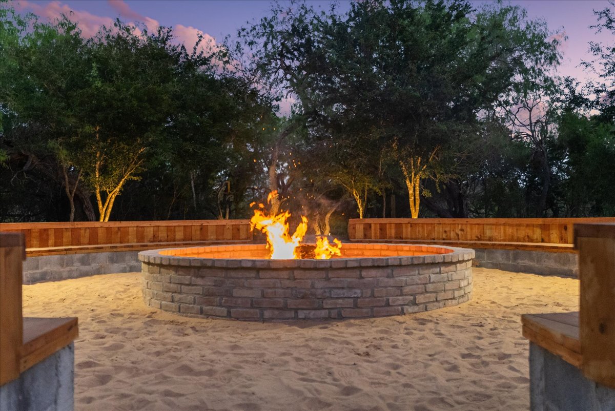 Circular brick fire pit with burning flames, surrounded by wooden benches on sandy ground, with trees and twilight sky in the background.