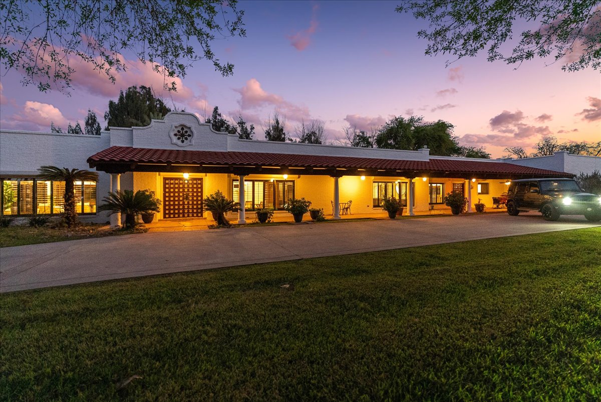 Single-story house with white walls and red tiled roof illuminated at dusk, with a parked black SUV and green lawn in front.