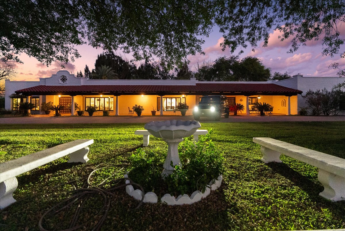 A white stucco house with red roof tiles illuminated at dusk, featuring a garden birdbath framed by two white benches on a grassy lawn.