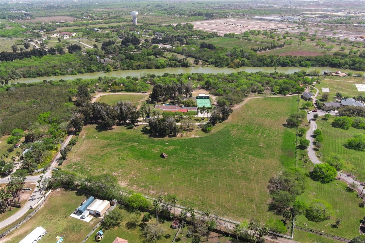 Aerial view of a large green field with scattered trees, a river running behind it, and several buildings and roads nearby in a rural area.